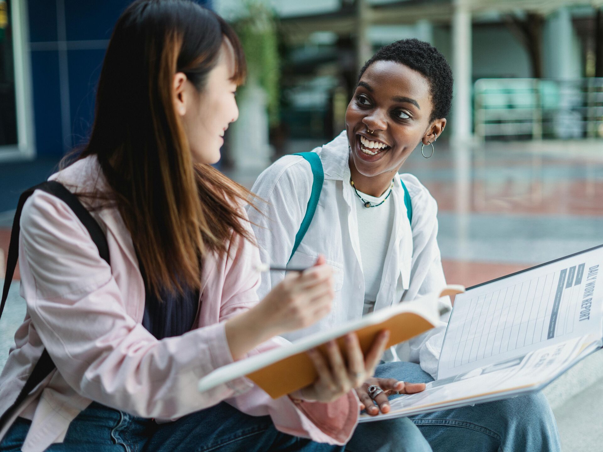 Zwei Studierende sitzen zusammen, unterhalten sich lachend und halten Notizbücher in den Händen.